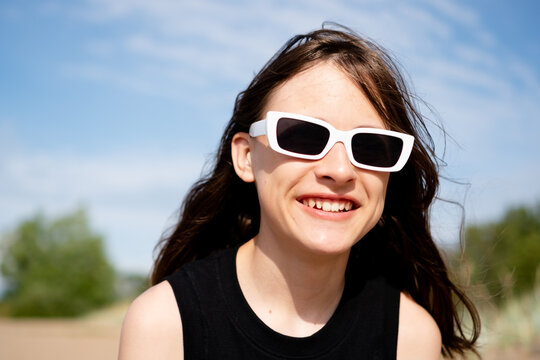 A teenage girl in white sunglasses beams with a joyful carefree smile on a sunlit beach, her hair dancing in the breeze. The teenager radiates pure happiness by the sea on a bright sunny day.