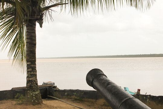 Shot of an antique iron cannon positioned on the grassy waterfront at Fort Nieuw Amsterdam in Suriname. This historic open-air museum sits at the confluence of the Suriname and Commewijne rivers