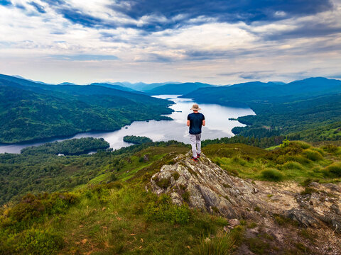 Solo hiker stands on the summit of Ben A'an mountain above Loch Katrine in Scotland, UK, with dramatic clouds, green hills and a calm lake stretching through the Highlands.