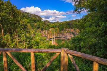 Fototapeta premium View of Chamarel Waterfall framed by a wooden railing and dense tropical forest in southwest Mauritius under a bright sky.