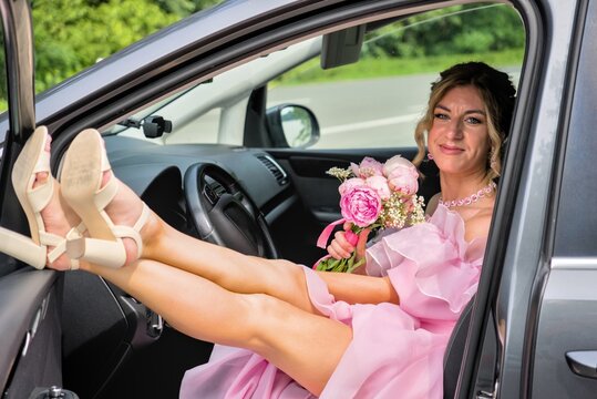 ​Happy beautiful woman in pink dress with peony bouquet and feet on car dashboard