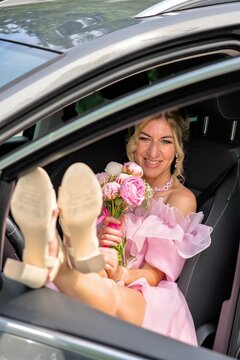 ​Happy beautiful woman in pink dress with peony bouquet and feet on car dashboard