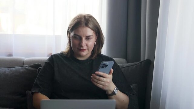 A woman is using a smartphone in a neutral living room setting, featuring a sofa and a window. The scene is captured from a fixed perspective, emphasizing daily life and technology themes.