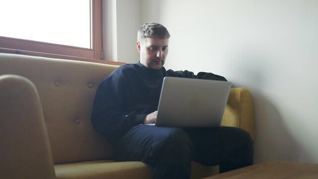 A side view of a young Caucasian man sitting on a modern yellow couch, holding his head thoughtfully while working on a laptop computer in a bright minimalist room.