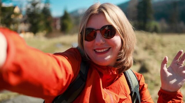 A close-up shot of a cheerful Caucasian female traveler in an orange jacket and sunglasses, smiling and taking a selfie photo during a mountain hike on a sunny summer day.