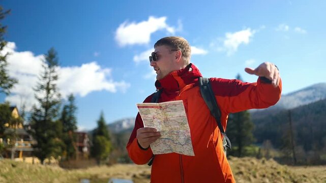 A young handsome male traveler in an orange jacket and sunglasses holding a paper map and pointing away on a sunny day during a mountain hike.