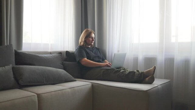 A wide shot of a young plus-size Caucasian woman sitting on a grey couch near a window, smiling at her phone screen while holding a laptop computer on her lap.