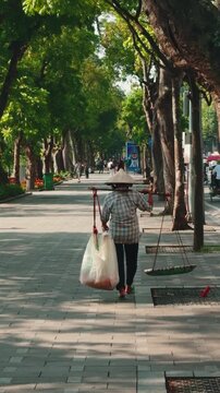 Vietnamese street vendor walking along a shaded promenade near hoan kiem lake. Carrying a plastic bag filled with items and a basket of produce using a traditional shoulder pole