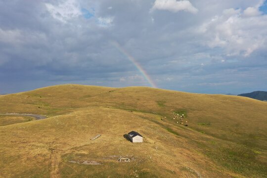 Aerial view of Campo Imperatore with a rainbow over rolling hills, a small stone building, and grazing cattle in L'Aquila, Abruzzo, Italy.
