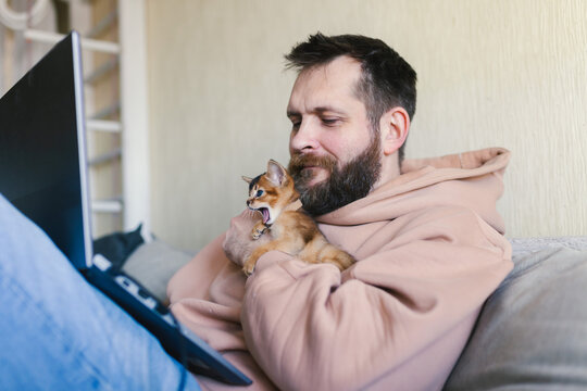 Bearded man working on laptop at home while holding and cuddling small yawning Abyssinian kitten. Work-life balance, remote work, companionship, cozy home atmosphere, love for pets.