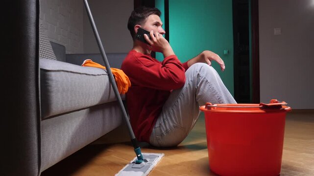 Young man sitting on the floor, calling using a mobile phone next to a mop and bucket, dealing with a leaked water problem in his home