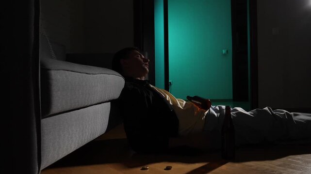 Man with an alcoholic drink bottle lying unconscious on the floor next to a sofa in a dark room, representing addiction, despair, and loneliness