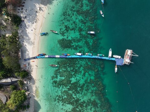 Aerial view of the floating pier at Ko Racha Yai with boats moored alongside, crystal clear turquoise water, and visible coral reefs near the sandy beach in Rawai, Phuket, Thailand.
