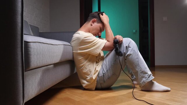 Frustrated young man sitting on the floor, holding a game controller and showing disappointment after an intense gaming session or losing a video game competition