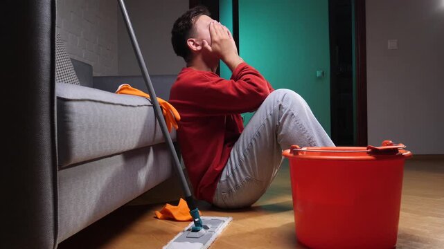 Young man sitting on the floor, covering his face with hands, feeling stressed and overwhelmed by home maintenance tasks after mopping the living room