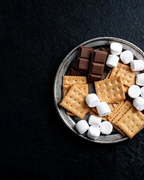 A vertical flat lay featuring dark chocolate bars, graham crackers, and marshmallows arranged on a round vintage metal plate over a dark slate background, leaving large copy space.