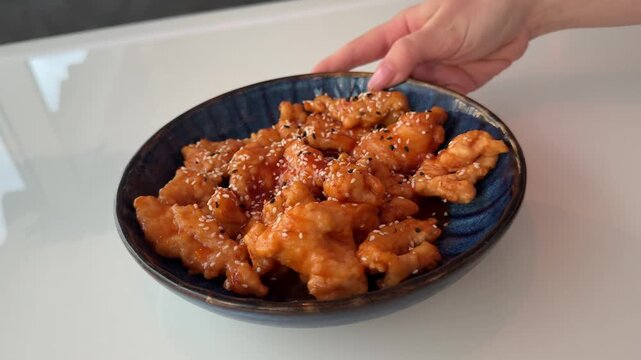 A girl gently rotates a blue plate filled with chicken wings in sweet and sour sauce with sesame, then lifts the dish from the white table