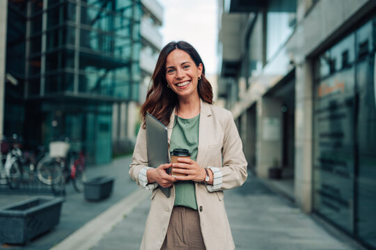 Smiling businesswoman holding laptop and coffee, walking in modern city, confident and successful career woman outdoors
