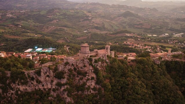 Aerial view of the historic Guaita Tower perched on rocky cliffs of Mount Titano, surrounded by lush hills and medieval architecture of Citta di San Marino, San Marino, San Marino.