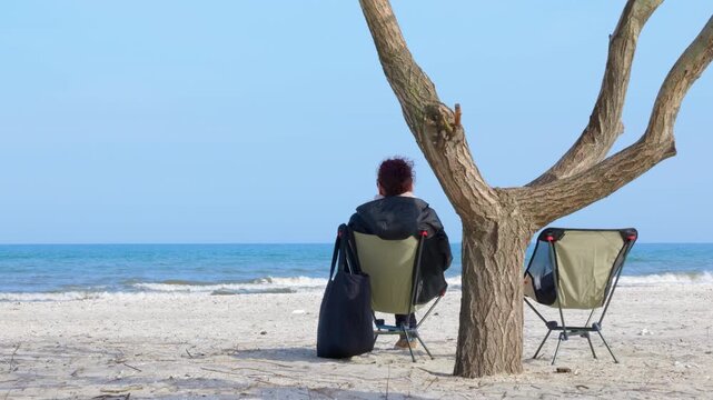 Woman enjoying the seascape. Solitary woman in a jacket sitting on a folding camping chair on a sandy beach, enjoying the peaceful view of the sea waves under a clear blue sky