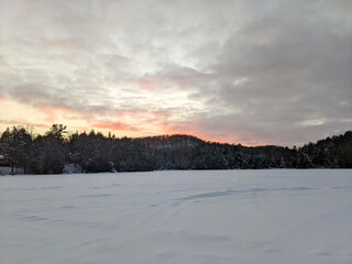 Naklejka premium A wide landscape shot of a vast frozen lake completely covered in white snow, bordered by a dense winter evergreen forest line under a dramatic cloudy sunset sky with orange hues.