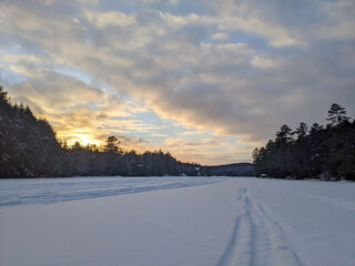 Naklejka premium A wide landscape shot of multiple snowmobile tracks intersecting across a vast frozen lake covered in deep white snow, with a dense forest line under a soft sunset sky with pastel clouds.