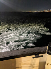 Obraz premium A shot from a boat looking down at a field of circular pancake ice sheets floating on dark water at night, with a brightly lit city skyline visible on the horizon.