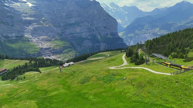 A wide, scenic aerial pan shot of a yellow and green passenger train descending along a hillside rack railway track, with a winding road, small mountain buildings, and grazing cattle below, against a 
