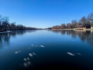 Obraz premium A wide angle view of a completely frozen lake with trapped air bubbles on Toronto Island. Bare winter trees line the snowless shore next to an empty park pavilion building on a bright sunny day.