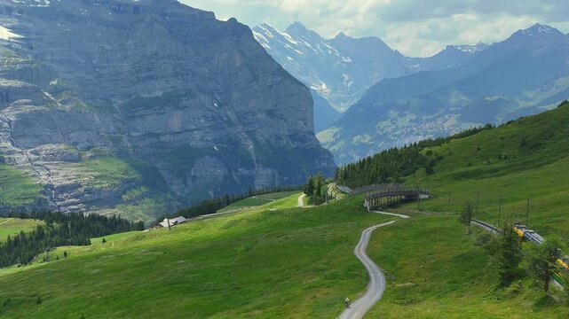 A high-angle aerial view of a yellow and green passenger train traveling on an alpine rack railway, passing alongside a winding gravel path with a cyclist, set against a majestic backdrop of massive r