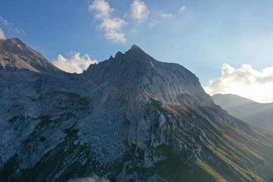 Aerial view of Corno Piccolo featuring rugged limestone peaks, rocky slopes, and grassy mountain ridges under a bright blue sky in Pietracamela, Abruzzo, Italy.
