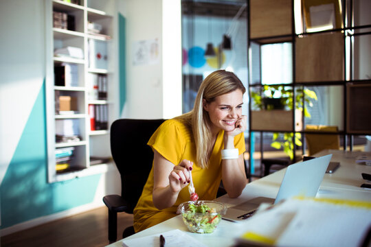 Smiling businesswoman eating salad while working on laptop in modern office