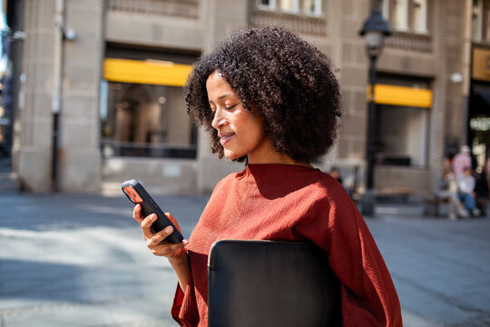 Young professional checking smartphone and holding laptop on city street