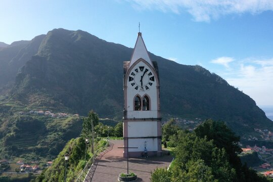 Aerial view of Capelinha de Nossa Senhora de Fatima featuring its distinctive clock tower set against lush green mountains in Sao Vicente, Madeira, Portugal.