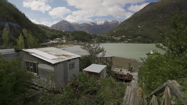 Landscape view of Caleta Tortel, Chile, featuring rustic houses with metal roofs, a smoking chimney, overlooking a bay surrounded by lush forest and snow-capped mountains.