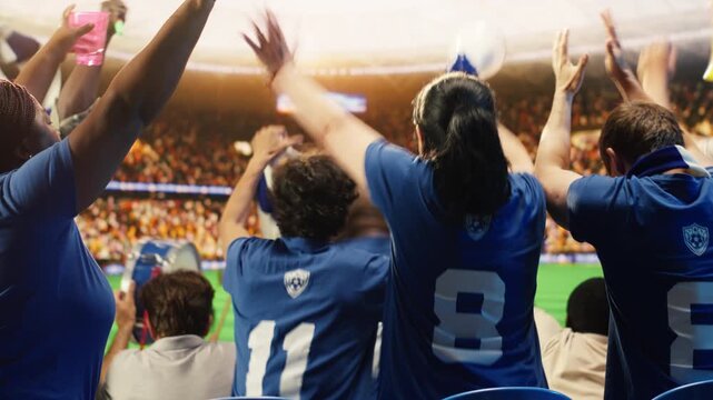 Rear view of happy football supporters in blue team jerseys raising fists to celebrate during soccer match day in packed stadium, showing soccer crowd jumping from joy after after competition victory.