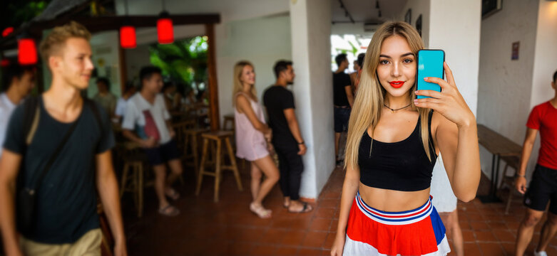 young blonde woman with a gentle smile holding a smartphone with a blank blue screen inside a casual tropical caf&eacute;