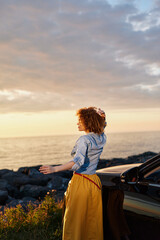Fototapeta premium Cheerful woman by the sea at sunset, wearing a blue shirt and yellow skirt, leaning against a car, enjoying breeze and warm light in a relaxed lifestyle moment outdoors
