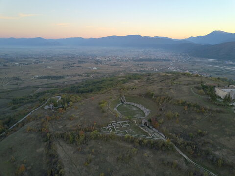 Aerial view of the ancient Roman amphitheater and archaeological ruins of Alba Fucens set in a hilly landscape at sunset in Albe, Abruzzo, Italy.