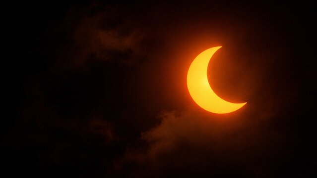Glowing orange partial solar eclipse crescent shining through dark dramatic clouds in the sky