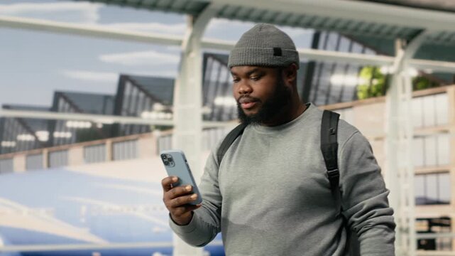 Happy man walks through airport terminal with smartphone and backpack, cheerful African American traveler using a mobile phone for texting in airport terminal, for aviation travel concepts.