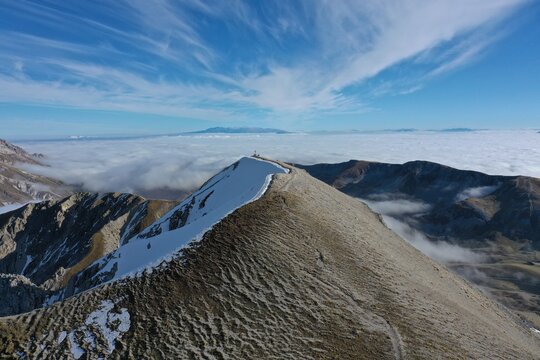 Aerial view of Monte Aquila with snow-dusted slopes and a sea of clouds below under a clear blue sky in L'Aquila, Abruzzo, Italy.