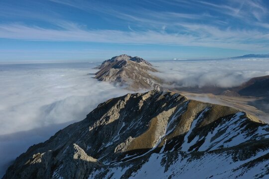 Aerial view of Monte Aquila rising above a thick sea of clouds with snow-dusted rocky peaks under a clear blue sky in L'Aquila, Abruzzo, Italy.