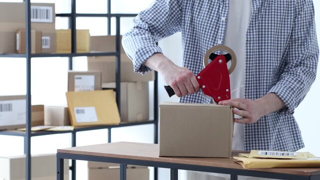 Man using dispenser with adhesive tape to seal cardboard box and sticking barcode onto it before sending at table in office, closeup