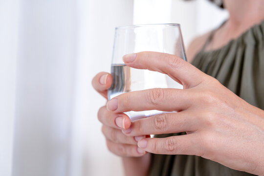 Woman in a green top holding a clear glass of fresh drinking water with both hands in front of a bright window at home