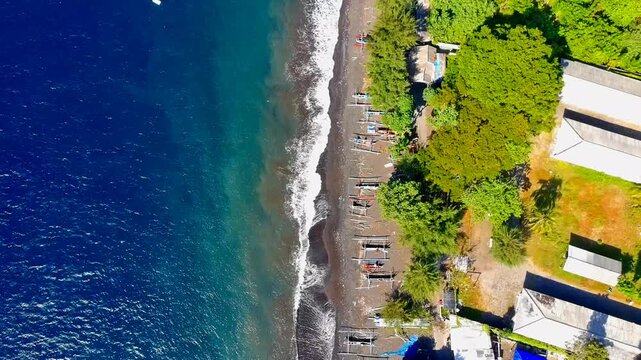 Aerial view of Pantai Waru Doyong Bulusan beach with traditional boats on the dark sand shore, lush trees, and coastal buildings in Kecamatan Kalipuro, Jawa Timur, Indonesia.