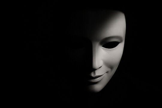 A white theatrical mask with a subtle smirk half-hidden in deep, dramatic shadow.