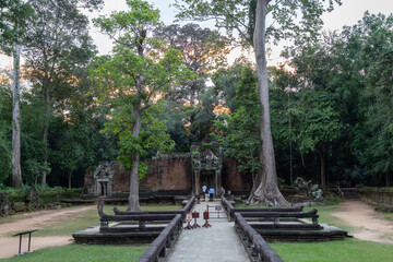 Obraz premium Two tourists walk towards the ancient temple ruins of Banteay Srei in Siem Reap, Cambodia, surrounded by lush jungle foliage.