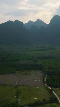 Vertical screen Aerial Approach Over Rice Plain And Rural Lane In Phong Nha With Karst Backdrop, Muddy Paddy Plots, Irrigation Channels And Scattered Village Huts Under Soft Sky