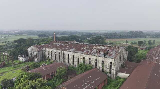 Drone footage pulling backward from Kushtia Sugar Mill, expanding the view to include factory buildings, industrial infrastructure, surrounding roads, and the nearby landscape.